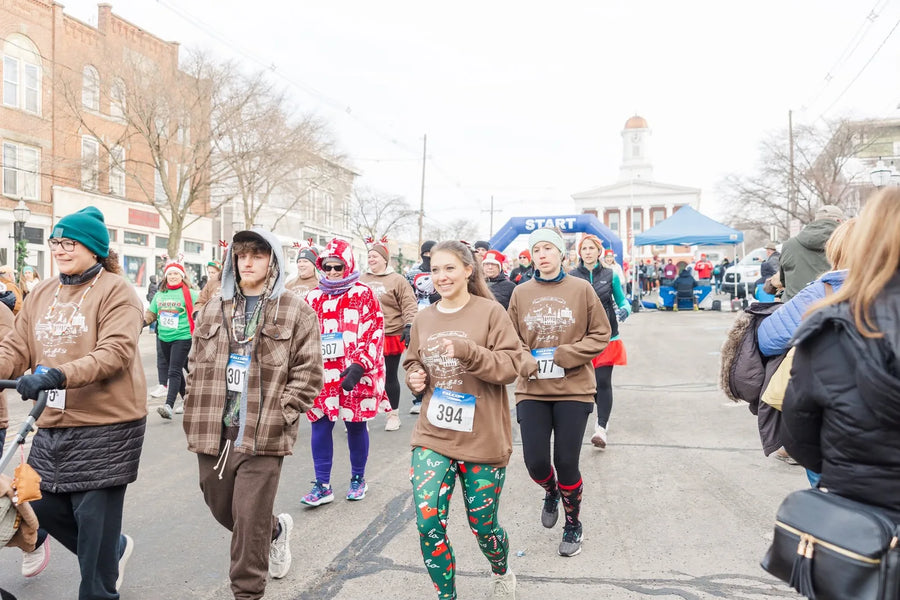 Runners and walkers in festive clothing on the Jingle Bell 5K course in downtown Montrose, Pennsylvania, with the start arch in the background.